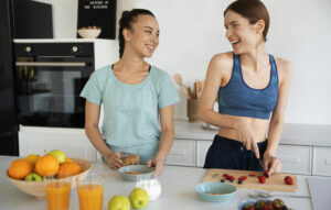 Deux jeunes femmes souriantes en cuisine, en train de préparer un petit-déjeuner sain avec des oranges, du jus de fruits et des aliments sans sucre raffiné, illustrant la question : faut-il supprimer totalement le sucre de son alimentation ?