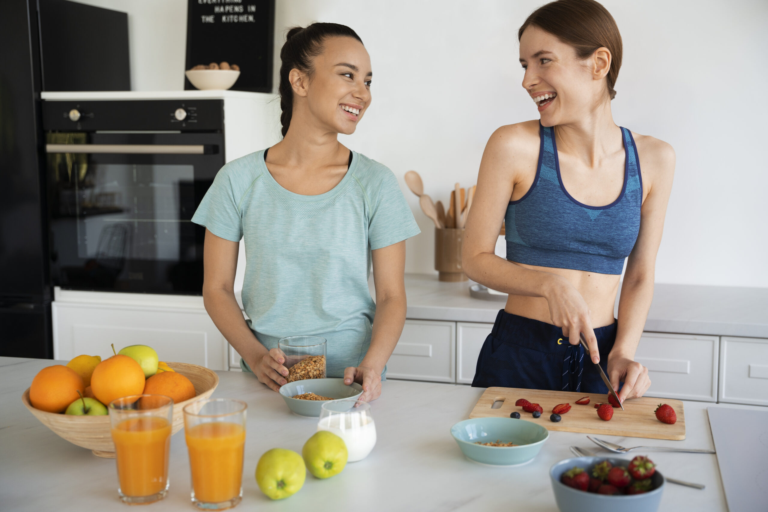 Deux jeunes femmes souriantes en cuisine, en train de préparer un petit-déjeuner sain avec des oranges, du jus de fruits et des aliments sans sucre raffiné, illustrant la question : faut-il supprimer totalement le sucre de son alimentation ?
